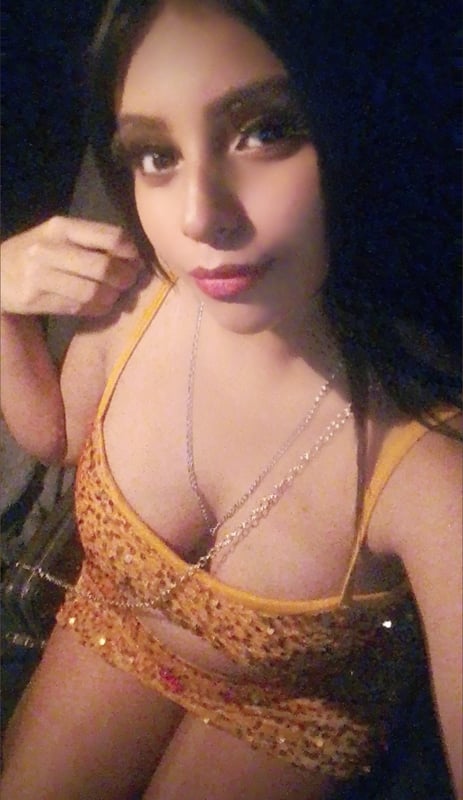 Young woman with long dark hair posing in a shiny yellow top and necklace in a dimly lit setting.