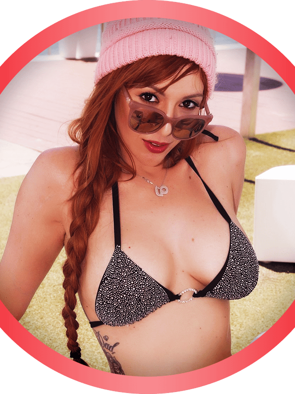 A woman with long red braided hair wearing sunglasses and a pink beanie, smiling in a black bikini top by a poolside.