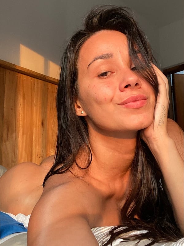 A woman taking a close-up selfie with a smile, lying down in a sunlit room with wooden walls.