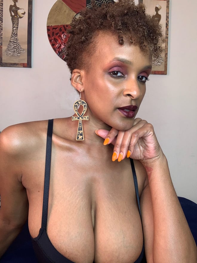 A woman with short curly red hair wearing a black top, adorned with African-inspired art and jewelry, posing thoughtfully indoors.