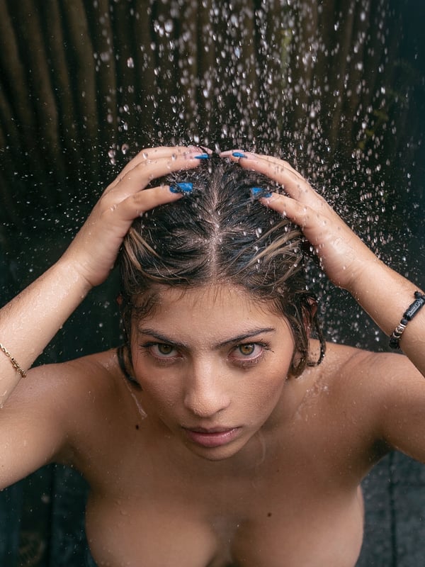 A woman with wet dark hair stands under an outdoor shower, hands running through her hair, with water streaming down in a natural bamboo enclosure.