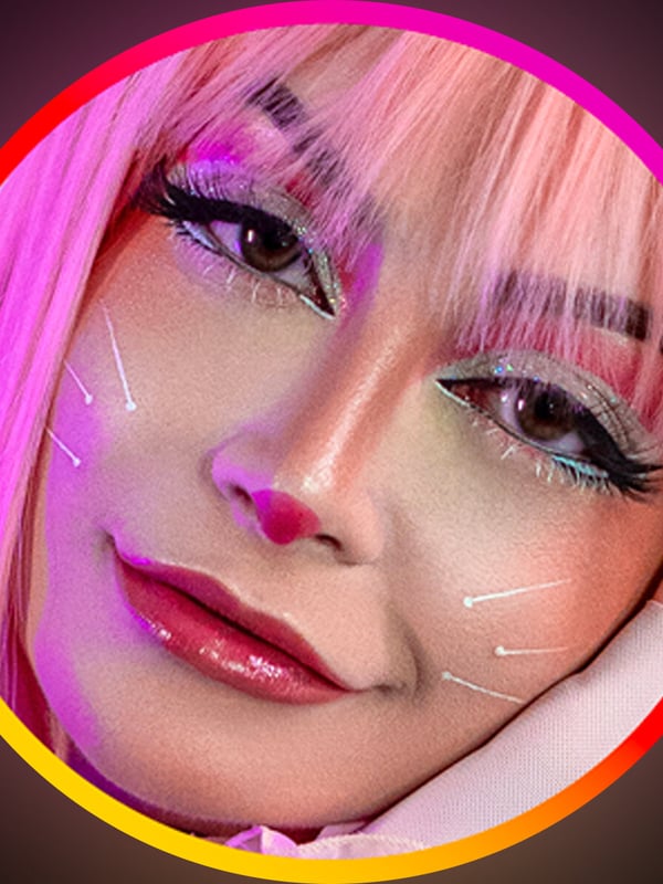 Close-up portrait of a young woman with vibrant pink hair, heavy makeup, and sparkling facial accents, framed in a glowing pink and purple circular border.