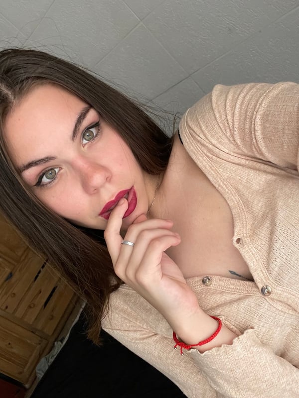 A young woman with long brown hair posing in a black tank top in a bedroom setting.