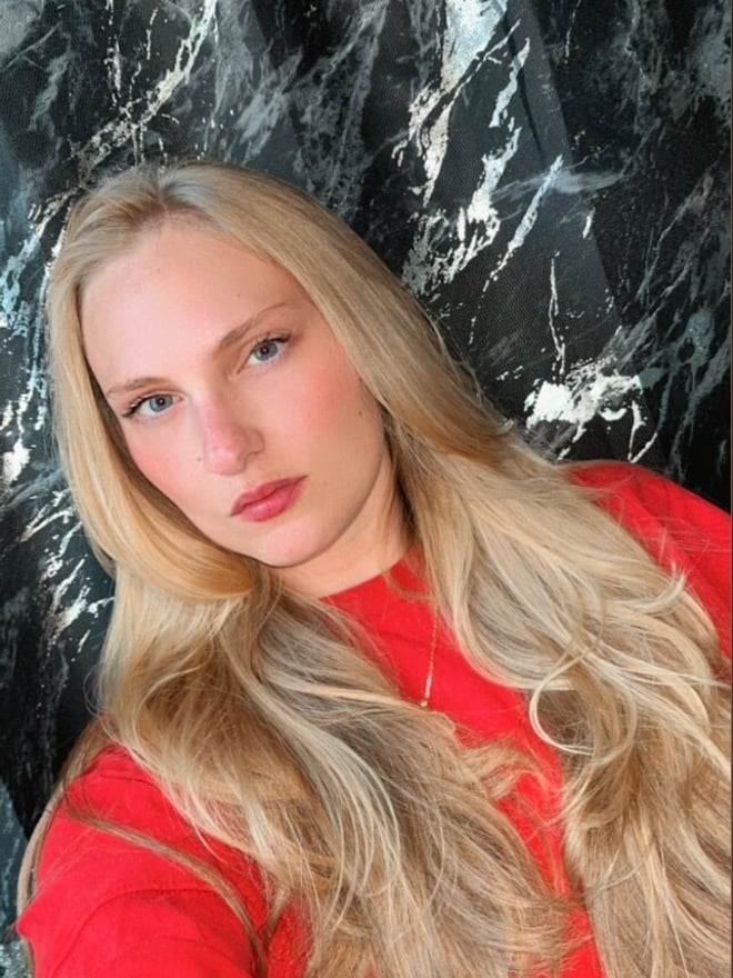 Young woman with long blonde hair wearing a red top, posing against a black marble background.
