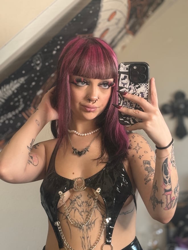 A tattooed person with dark magenta hair and bangs posing indoors in patterned clothing, with colorful ambient lighting in the background.