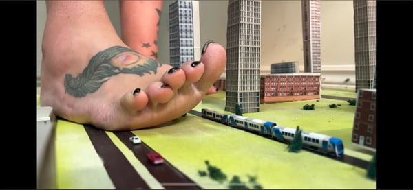A large foot positioned over a detailed miniature cityscape with tiny buildings, vehicles, and a train on a green base.