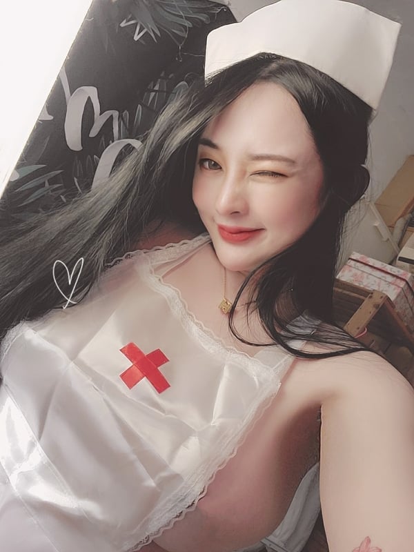 A woman dressed in a nurse costume with a white cap, smiling playfully while winking at the camera.