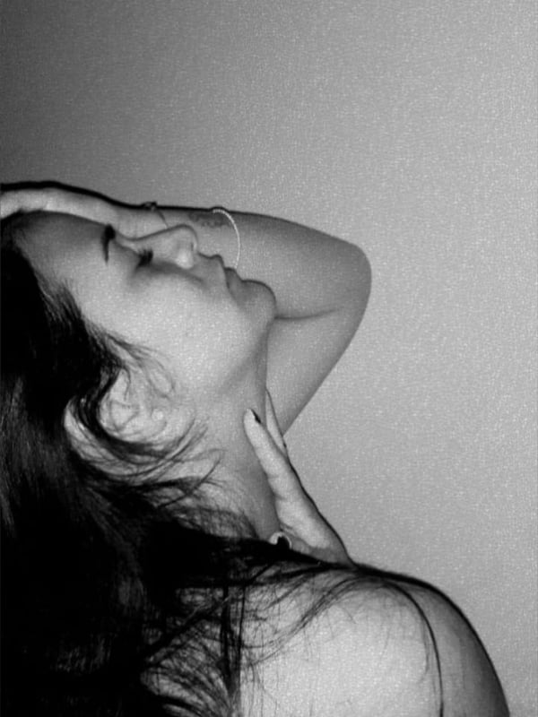 A black-and-white close-up of a woman with long dark hair, hand gently touching her neck, gazing sideways.