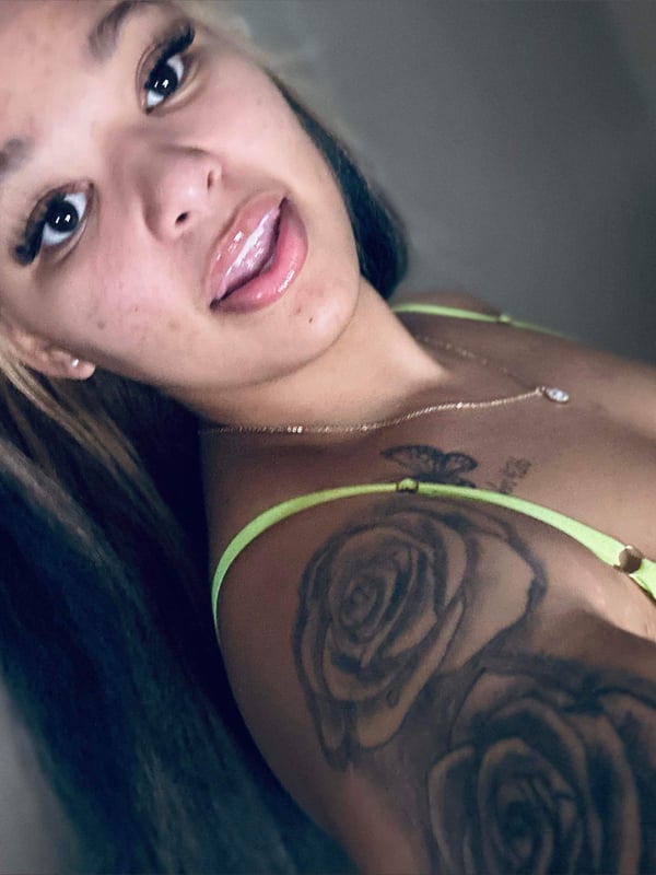 A young woman with blonde and dark hair in pigtails, wearing a green lace top, posing confidently indoors with visible tattoos on her arm.