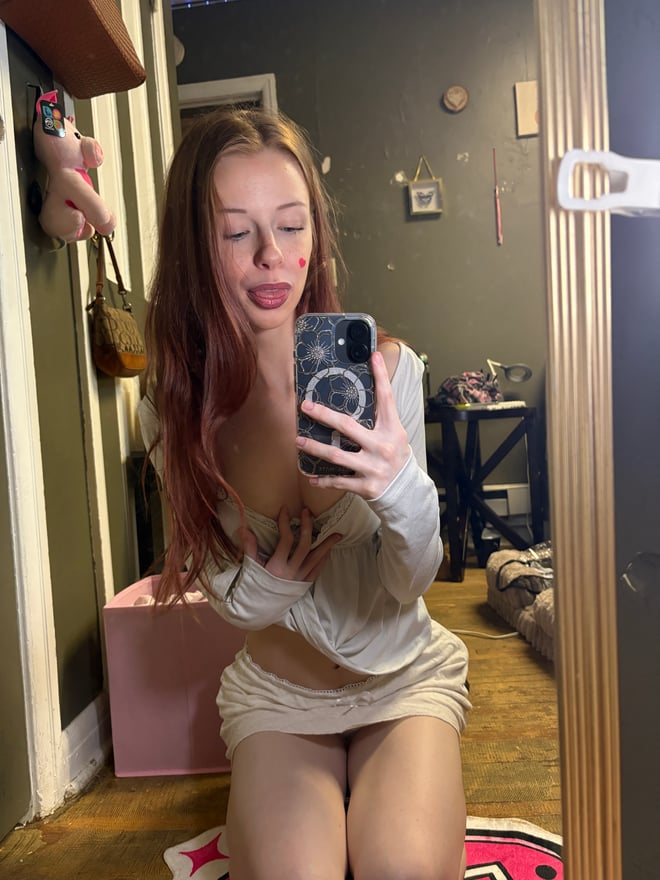 A young woman taking a mirror selfie while holding a pink plush toy, dressed casually in a bedroom.