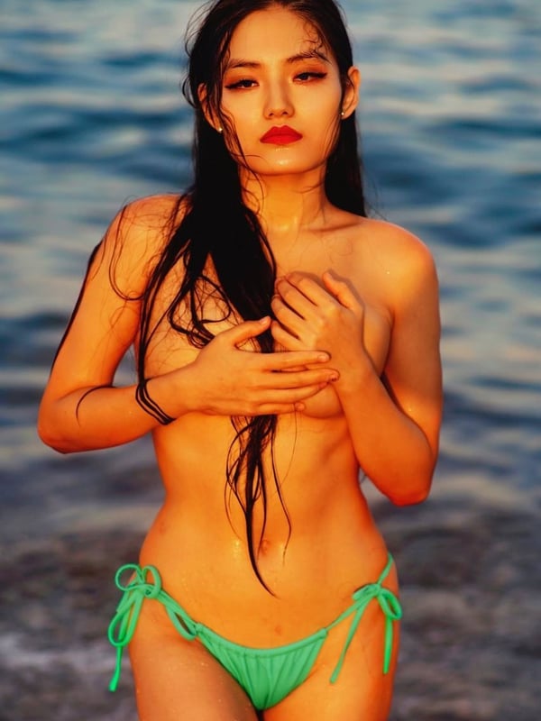 A young woman standing topless in a green bikini bottom by the ocean with wet hair.