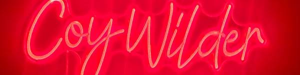 Neon pink sign displaying the name 'Coy Wilder' in cursive font against a dark background.