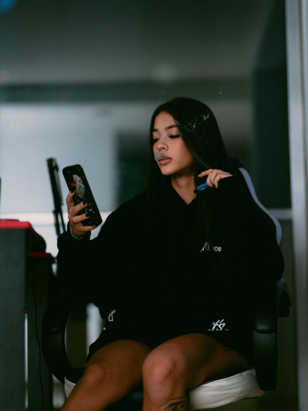 Young woman with long dark hair sitting in a black hoodie, holding phones, with tattoos on her leg, against a mirrored background.