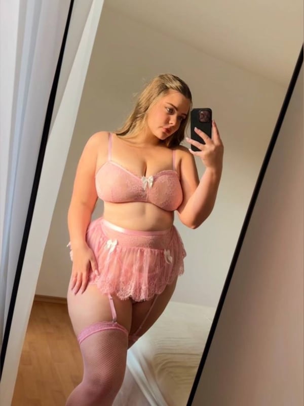 A woman standing in front of a mirror, wearing a pink lace lingerie set with garters and stockings, taking a selfie.