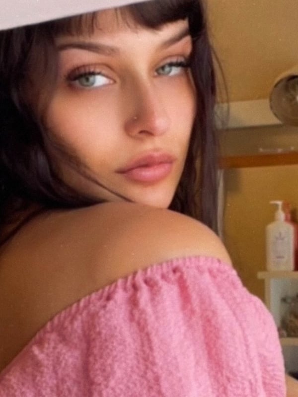 Close-up selfie of a young woman with long dark hair, green eyes, and pink lips, wearing a pink off-shoulder top, posing in front of a bathroom shelf with beauty products.