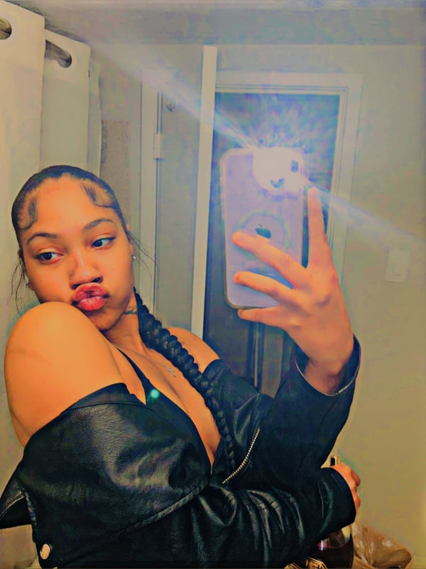 A young woman with braided hair taking a selfie in a room, wearing a black off-shoulder top and holding her phone.
