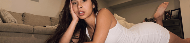 A young woman with long dark hair posing indoors on a bed in a white outfit.