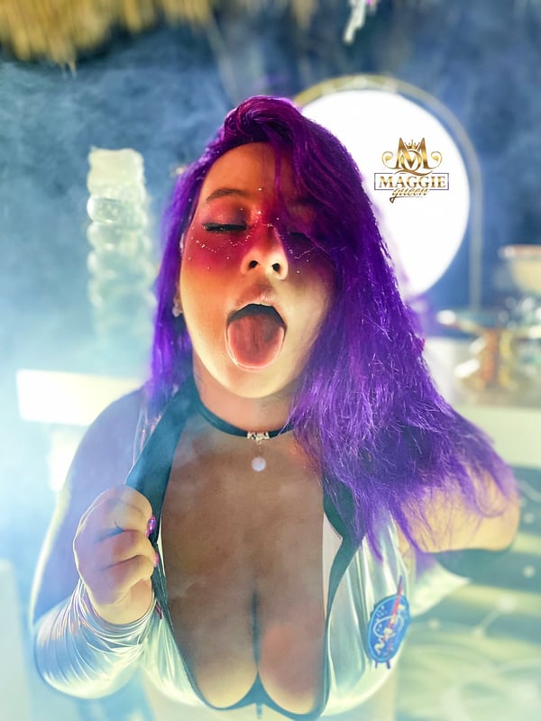 A woman with long purple hair posing playfully in a dimly lit, artistic setting with tropical elements and soft lighting.