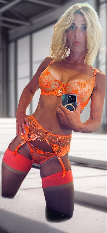 A woman with blonde hair posing confidently in ornate lingerie against an industrial warehouse background.