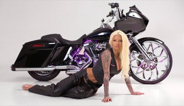 A woman with long blonde hair and tattoos poses on the ground next to a customized purple motorcycle in a studio setting.