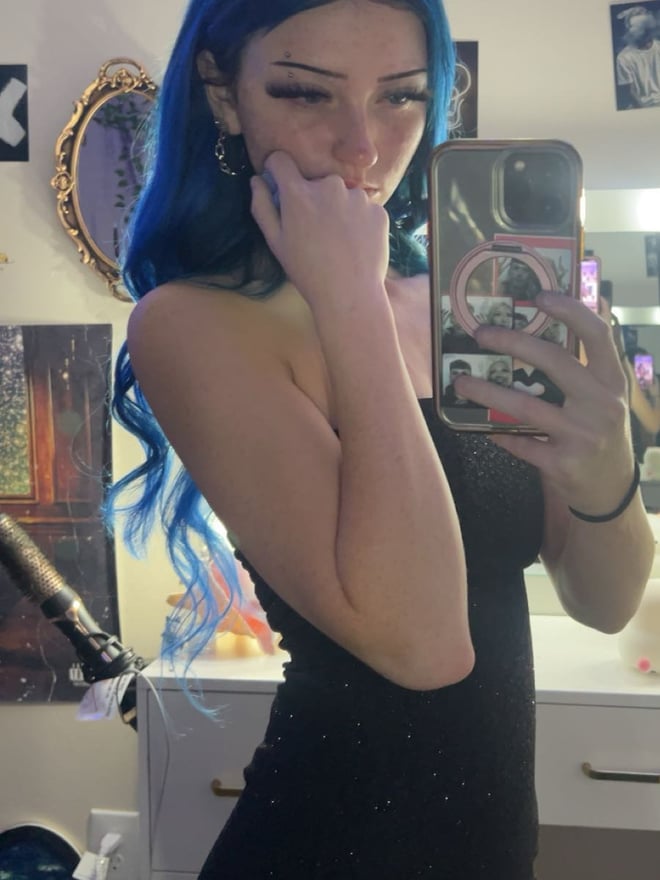 A young woman with long blue hair posing for a mirror selfie in a checked top.