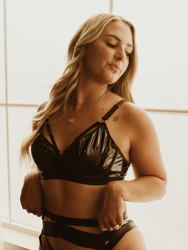 A woman with long blonde hair stands by a window in shiny black lingerie, smiling softly.