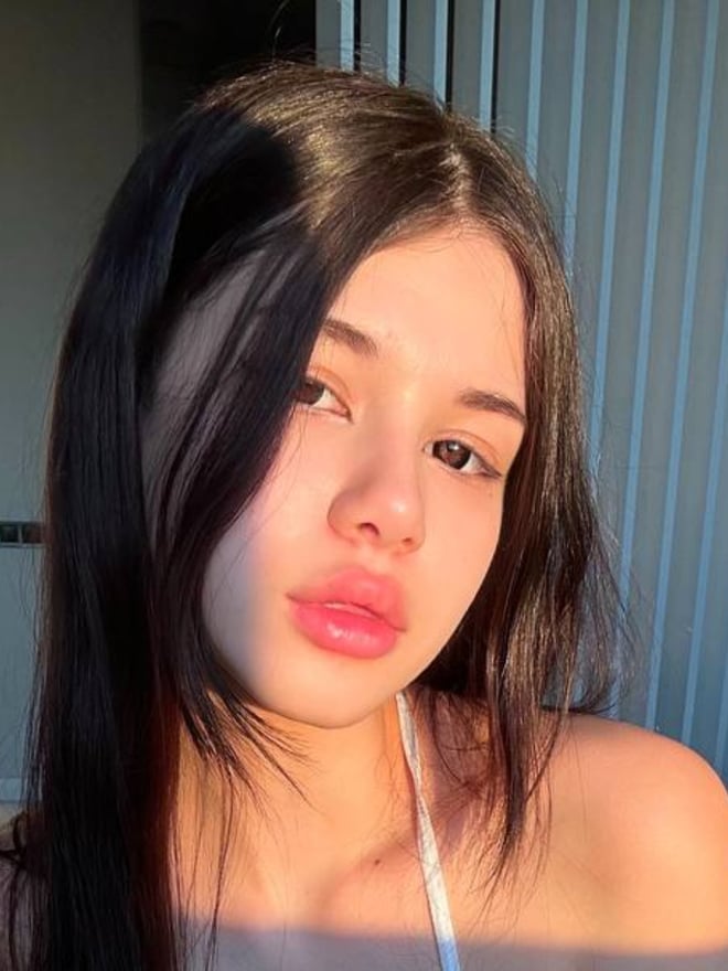 A young woman with long dark hair posing with glossy lips and a subtle smile in a sunlit indoor setting.