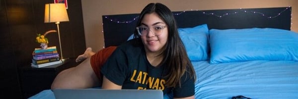 A woman with glasses and long brown hair lying on a bed in a cozy bedroom, wearing a casual t-shirt and smiling at the camera.