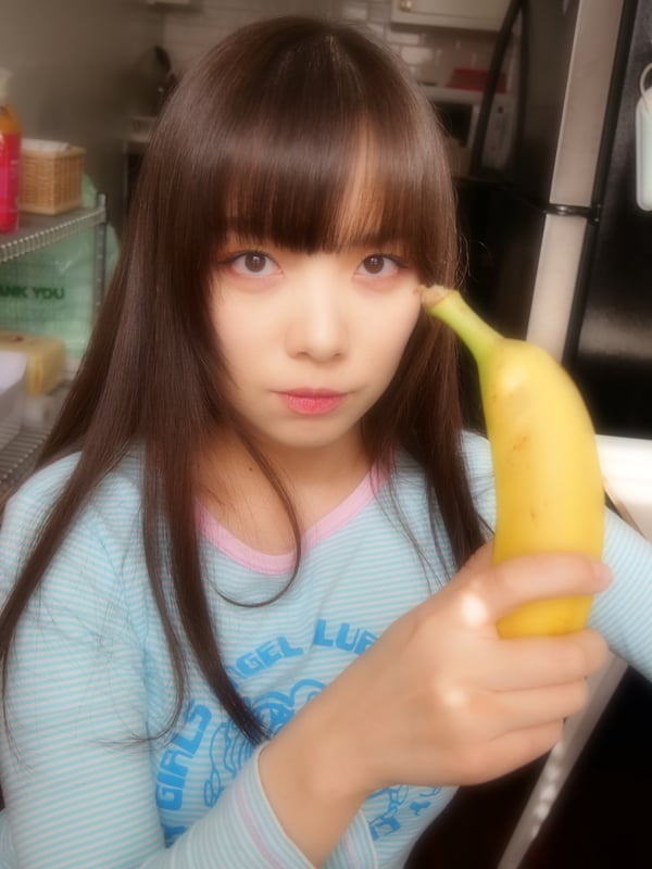 A young woman with long dark hair wearing a light blue sweater, holding a banana in a kitchen setting with shelves and plants.
