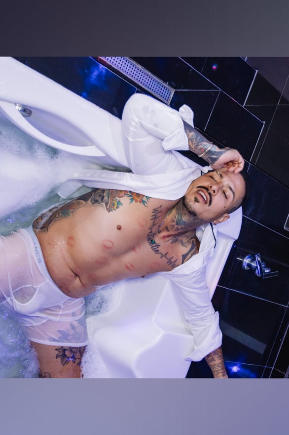 Man reclining in a bubble-filled bathtub, wearing an open white shirt and underwear, with tattoos visible on his arms and torso.