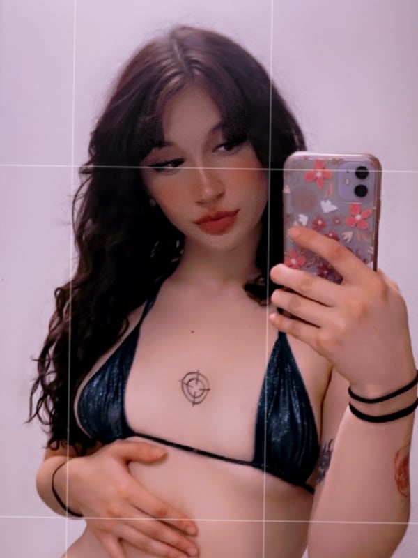 Young woman taking a mirror selfie in a black bikini, holding her phone with a pink case.