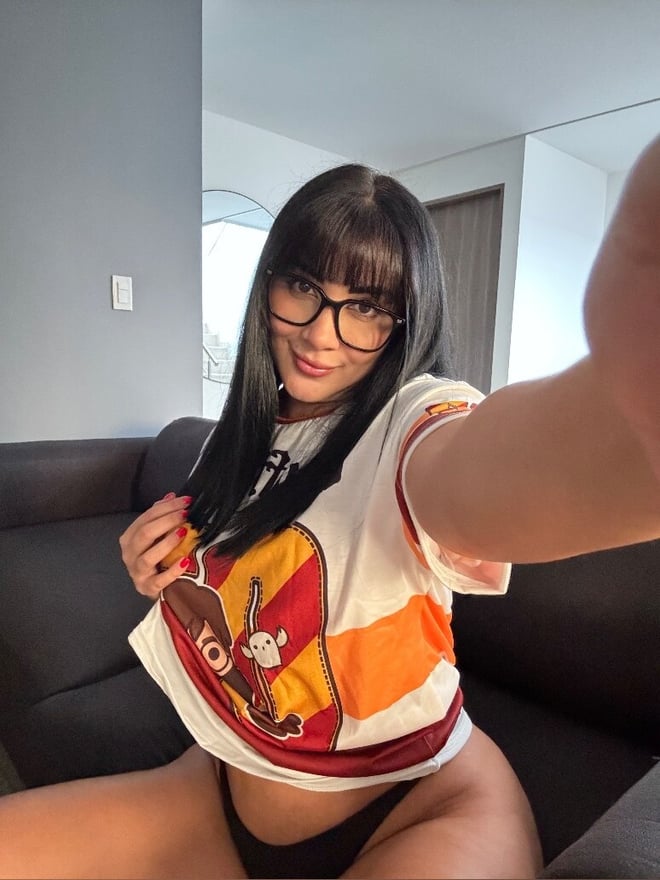 A young woman with glasses and long dark hair wearing a colorful striped shirt, smiling at the camera in a close-up selfie on a couch indoors.