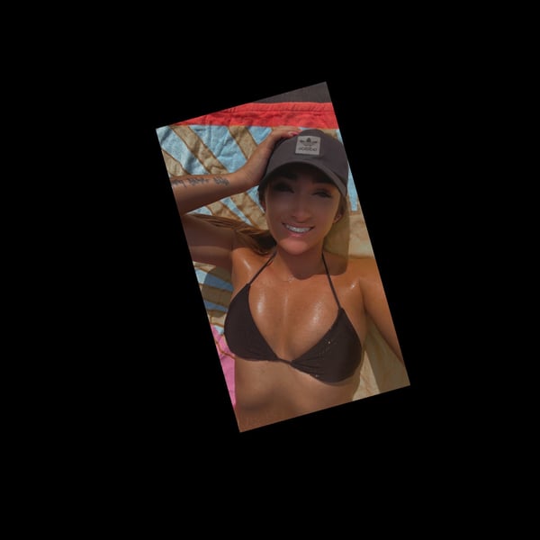 A woman smiling in a black bikini top and cap, lounging on colorful beach towels outdoors.