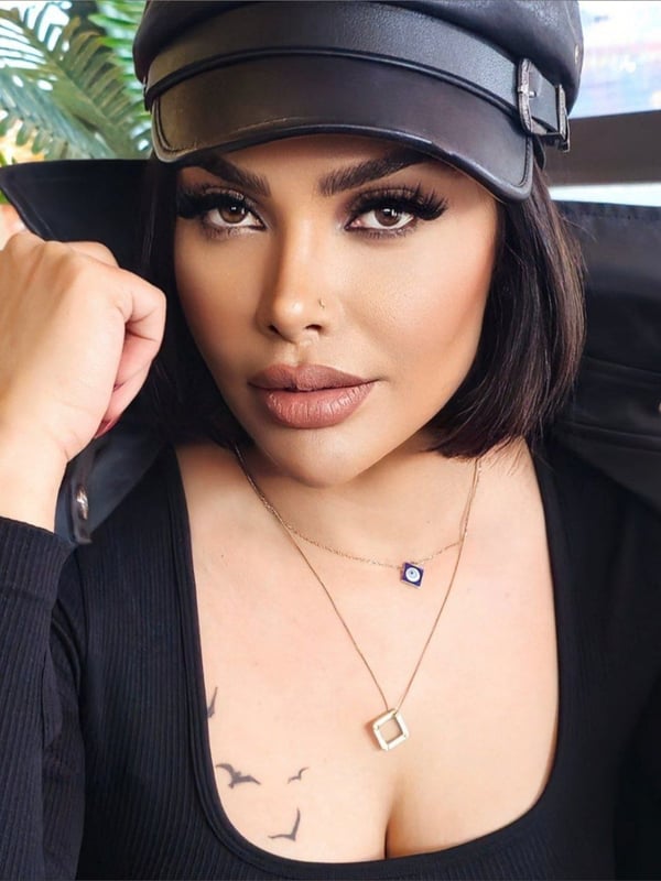 Close-up portrait of a woman wearing a leather hat and jacket, posing with bold makeup and jewelry.