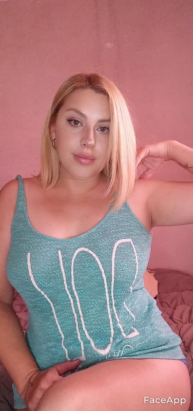 A woman with blonde hair posing on a bed in a teal tank top against a pink background.