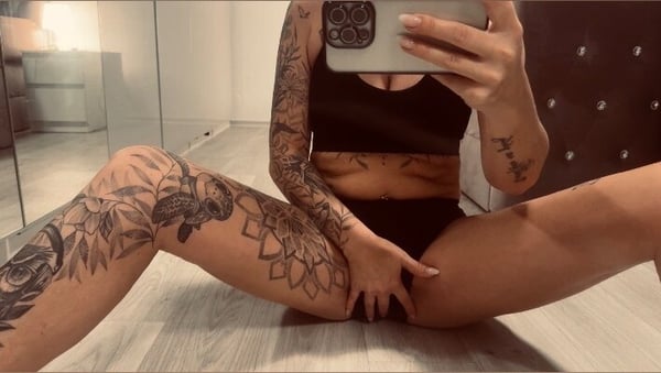 A woman with extensive tattoos sits on the floor in a black top and bottoms, holding a phone for a mirror selfie in a modern bedroom.