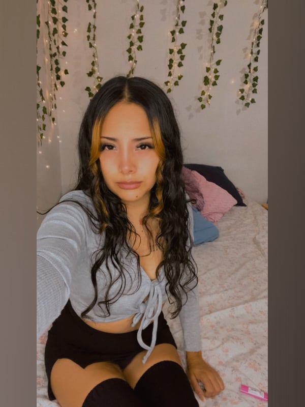 Young woman with long wavy dark hair highlighted in blonde, wearing a gray cropped top and black shorts with thigh-high stockings, posing on a bed with plant decor and soft lighting.