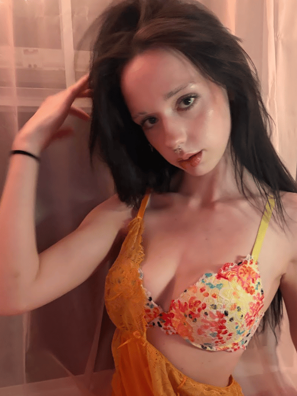 A woman posing in a colorful floral bra and sheer orange outfit, with her hand in her hair.