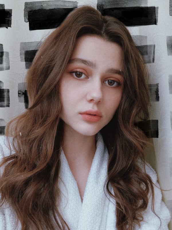 Young woman with long wavy brown hair wearing a white robe, posing against a black and white striped background.