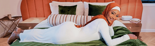 A woman in a white outfit and brown headscarf reclining on a bed in a warmly decorated room.