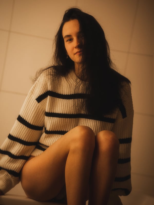 A young woman sitting with legs visible, wearing a striped sweater in a warmly lit tiled room.