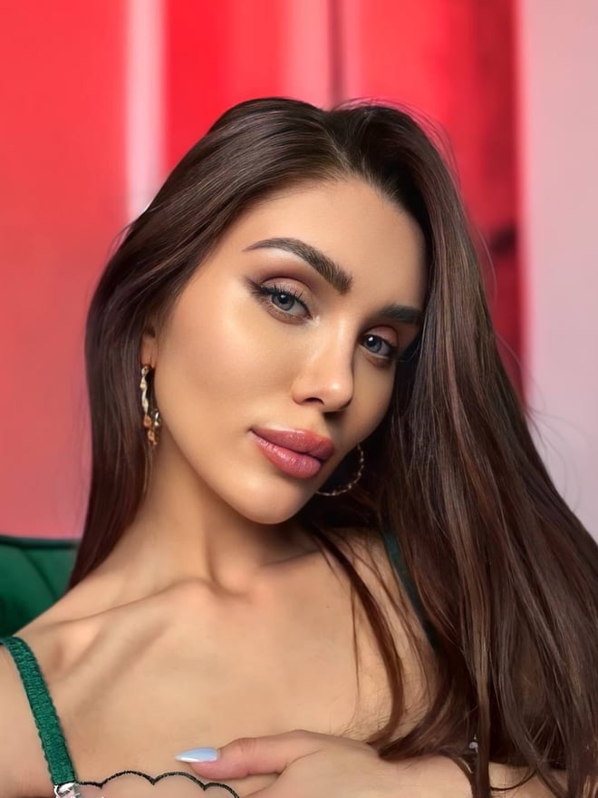Close-up portrait of a woman with long brown hair wearing a green top and earrings against a red background.