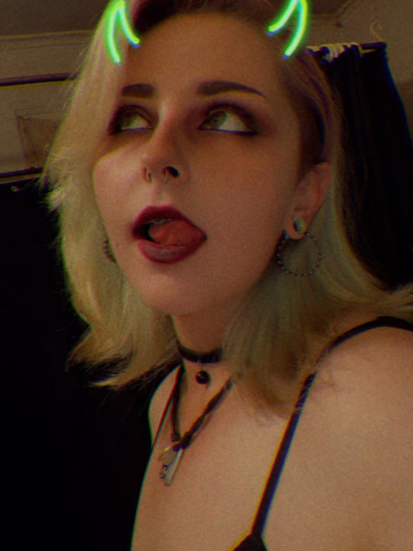Close-up portrait of a woman with platinum blonde hair, green glowing horns effect, and choker necklace against a dark background.