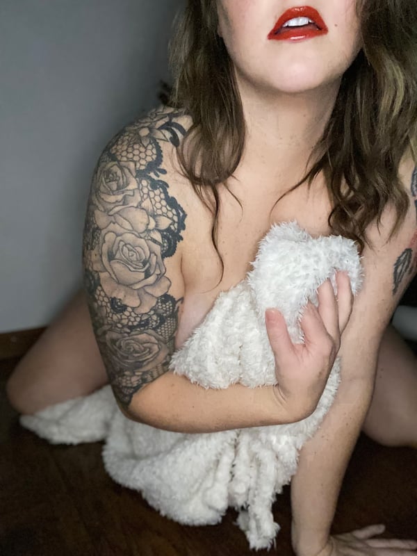Woman with blonde hair and tattoos sitting on the floor, holding a white fluffy fabric over her chest, smiling with red lipstick.