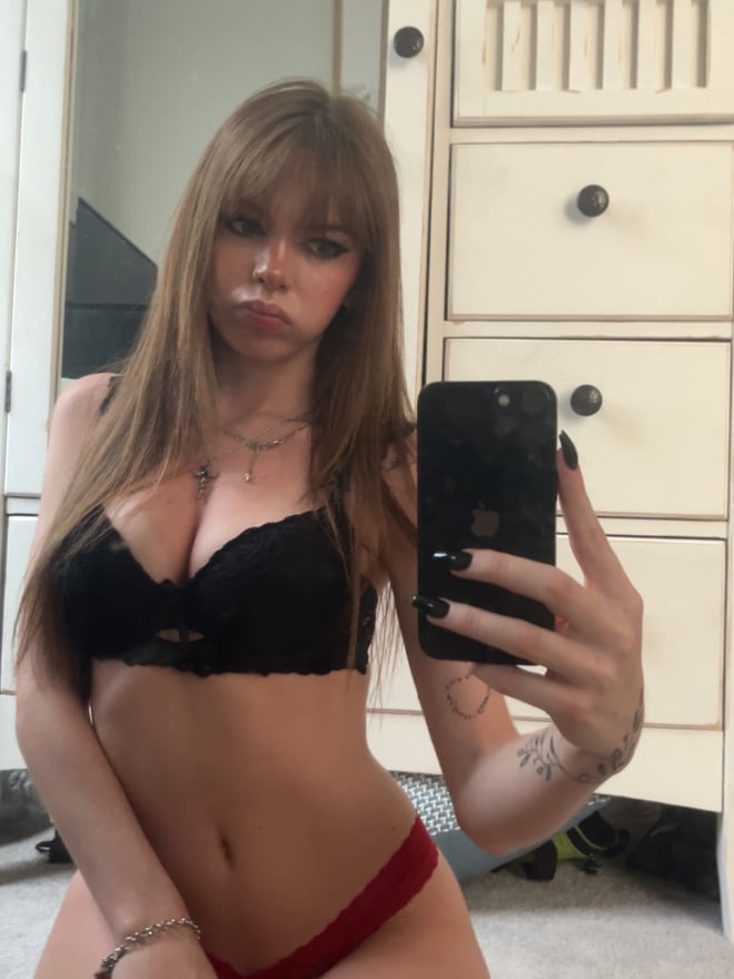 A person taking a mirror selfie wearing a black dress, with long brown hair and visible tattoos on the arm.