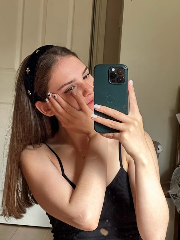 A young woman taking a close-up selfie indoors, wearing a black spaghetti strap top and headband, making a gentle hand gesture on her face.