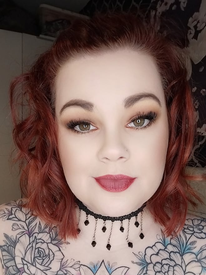 Close-up portrait of a woman with wavy red hair, green eyes, and makeup, wearing a beaded choker necklace against a dark patterned background.