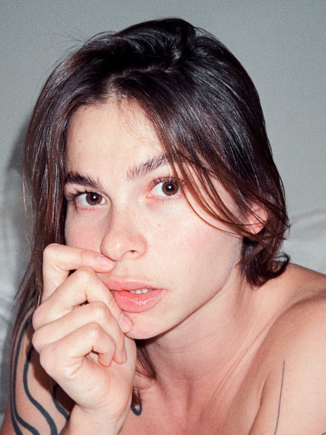 Close-up portrait of a woman with brunette hair, tattoos on her arms, and a subtle hand gesture near her face.