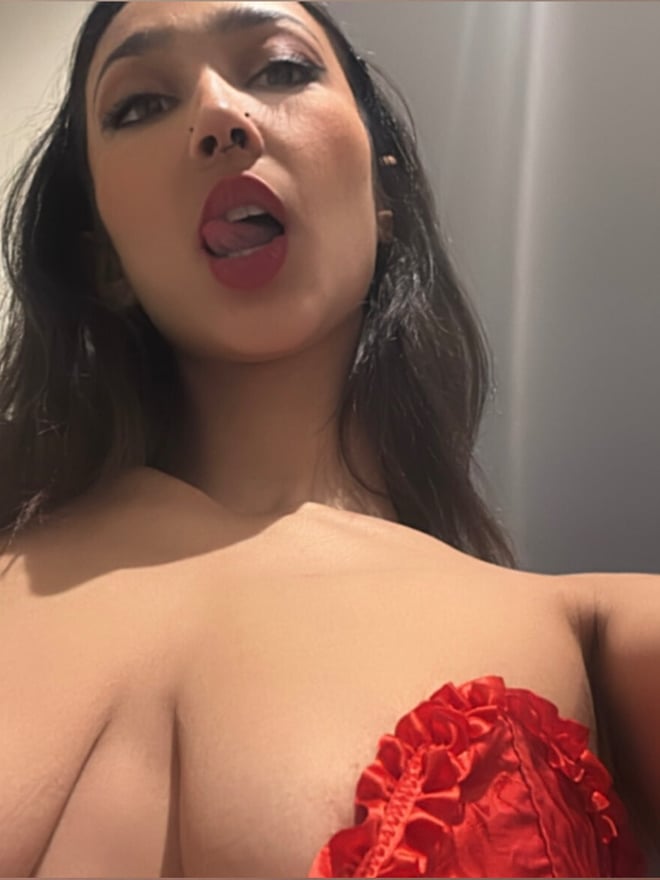 A woman with long dark hair making a playful face in a bathroom mirror selfie, wearing a red frilly top.