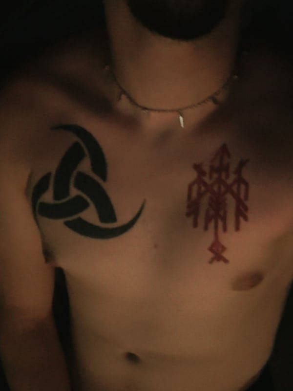Close-up selfie of a bearded man with extensive chest tattoos including Norse symbols, wearing a necklace, in low light.